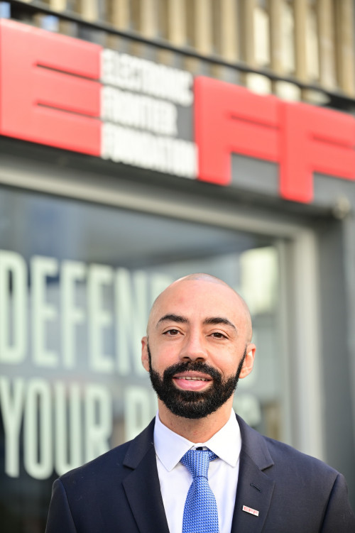 Head and shoulder image of nash in a blue suit jacket, white shirt and light blue tie, in front of building with EFF logo.