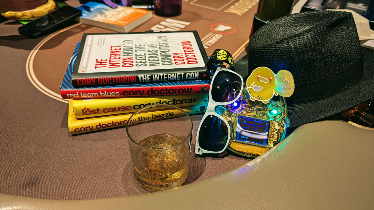 Photo of Cory Doctorow's Books, a drink, hat, and DEF CON 32 badge